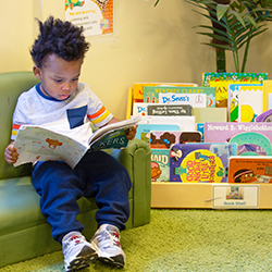 Toddler sitting on classroom floor looking at a book near a colorful learning toy shelf.